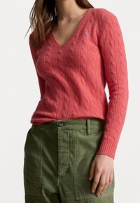 Sweter z dzianiny w kolorze koralowym z dekoltem w serek, z długimi rękawami i małym logo jako akcentem. Połączony z zielonymi spodniami typu cargo.