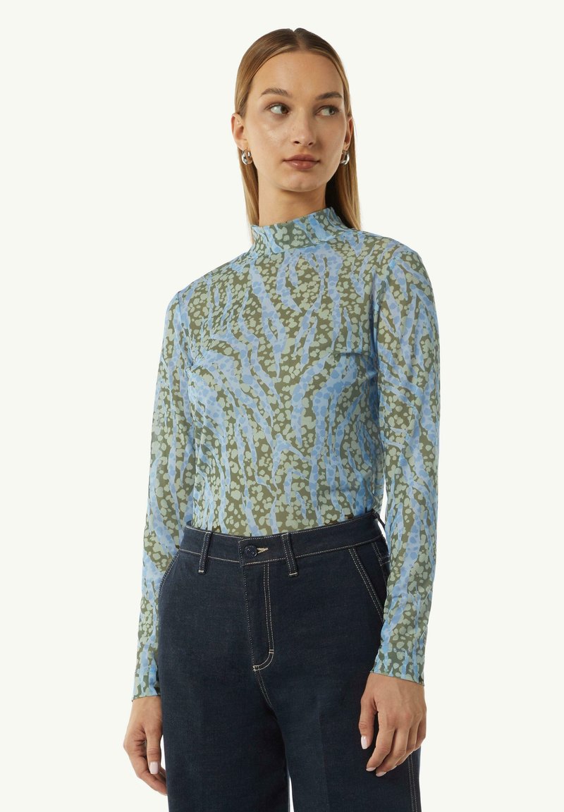 Langsleeve top in een lichtblauwe en groene abstracte print, met een hoge hals en een getailleerde pasvorm, gecombineerd met een donkere jeans.