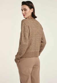 Brązowy sweter dziergany z teksturowanym wzorem w warkocze, ściśle przylegającymi mankietami i dołem oraz luźnym krojem. Połączony z beżowymi spodniami.