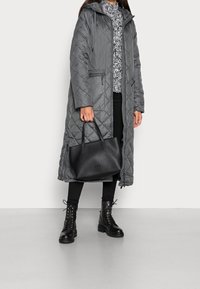Donna che indossa un lungo cappotto grigio trapuntato, leggings neri, stivali neri con lacci, mentre tiene una borsa a tracolla in pelle nera.