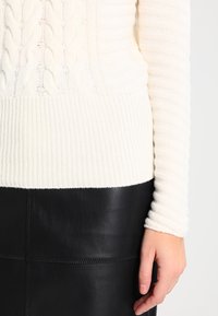 Weißer Kabelstrickpullover mit gerippten Bündchen und Saum, kombiniert mit einem schwarzen Lederrock mit glatter Textur und sichtbaren Nähten.