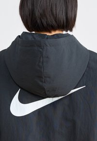 Melnā kapuce ar izteiktu baltu Nike swoosh logo aizmugurē, ar gludu tekstūru un pieguļošu kapuces dizainu.