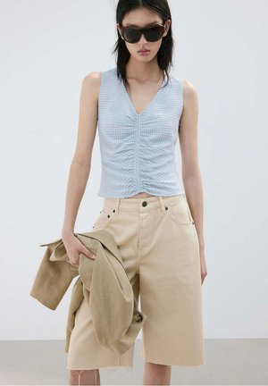 Femme portant un haut sans manches bleu clair froncé, un short beige longueur genou, des lunettes de soleil foncées, tenant une veste beige sur fond uni.