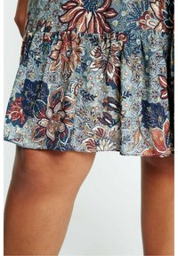 Gros plan sur une jupe courte à motif floral avec des fleurs bleues, rouges et beiges portée au-dessus des genoux sur des jambes bronzées.