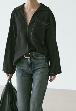 Person iført en sort langærmet skjorte med hvide syninger, blå jeans og holder en sort taske mod en ensfarvet lys baggrund.