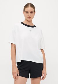 Frau mit zurückgegeltem Haar, die ein weißes, oversized T-Shirt mit schwarzem Kragen und schwarze Shorts trägt, steht vor einem weißen Hintergrund.
