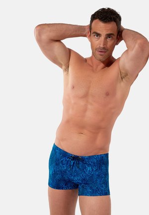 Athletischer Mann posiert mit den Händen hinter dem Kopf, trägt blaue Badeshorts mit dunklen Korallenriffmustern vor einfachem Hintergrund.