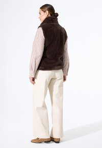 Braune Kunstfaserweste mit hohem Kragen, getragen über einem gestreiften Langarmhemd, kombiniert mit cremfarbenen weiten Hosen und hellbraunen Schuhen.