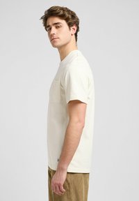 Cream short-sleeve t-shirt with a pocket on the left side, made of soft cotton material; worn with textured, ribbed brown corduroy pants.