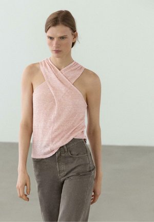 Kvinde iført en ærmeløs pink cross-wrap top og grå højhælede bukser, stående mod en ensfarvet lys baggrund.