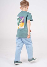 Grünes Baumwoll-T-Shirt mit dem 'BOFR'-Grafik in Lila, Gelb und Grün auf der Rückseite. Kombiniert mit hellblauen, lässigen Jeans und grünen Schuhen.