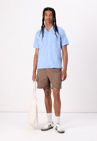 Chemise à manches courtes bleu clair avec des boutons, shorts marron, chaussettes blanches et baskets. Tient un sac fourre-tout crème. Texture lisse.