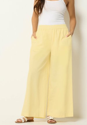 Frau trägt weißen Tanktop, weite gelbe Hosen mit elastischem Bund und Taschen und weiße flache Sandalen, steht vor einer schlichten Wand.