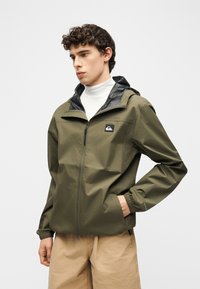 Νέος άνδρας που φοράει χακί ζακέτα με φερμουάρ και κουκούλα, μπεζ παντελόνι και λευκό ζιβάγκο, στέκεται με το ένα χέρι στην τσέπη.