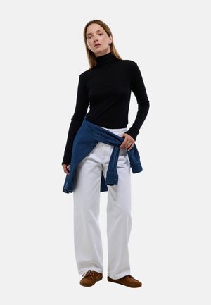 Un pull noir à col roulé, un pantalon large blanc et une chemise en denim bleu nouée autour de la taille. Des chaussures en daim marron complètent le look.