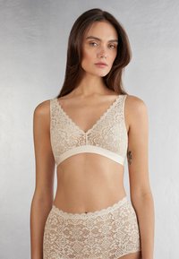 Krajkový braletka v nude barvě s vlnitými okraji, designem do V a širokými ramínky. Vytvořena v páru s odpovídajícími krajkovými kalhotkami s květinovými vzory.