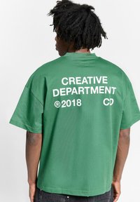 Camiseta verde de gran tamaño con el texto blanco "DEPARTAMENTO CREATIVO" y "2018 CD" impreso en la parte de atrás. Material de algodón suave, corte relajado.