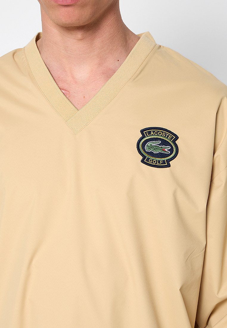 Polo beige à manches courtes avec un design en V. Présente un logo "Lacoste Golf" brodé en noir et vert foncé sur le côté gauche.