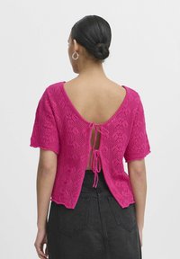 Rozā adīta blūze ar īsām piedurknēm un dziļu V-veida aizmuguri, ar mežģīņu rakstu un regulējamu saiti jostas vietā.