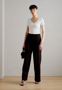 T-shirt blanc à manches courtes et col en V, pantalon noir taille haute avec plis, sac à main noir et sandales à lanières sur un sol en bois.