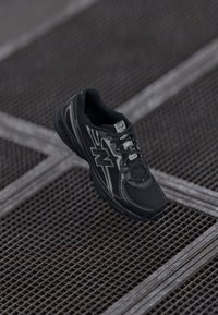 Baskets de sport noires avec une tige en maille texturée, des superpositions synthétiques, un col rembourré et une semelle extérieure en caoutchouc. Présente des accents argentés contrastés.