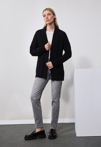 Schwarzer Strickcardigan mit offenem Vorderteil, strukturiertem Design, kombiniert mit hellgrauen Jeans und schwarzen Slippern, vor einem minimalistischen Hintergrund.
