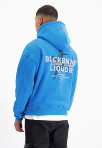 Blauwe hoodie met een grafische print op de rug. Heeft een geribde zoom en boorden. Draagt over een wit shirt. Zwarte broek.