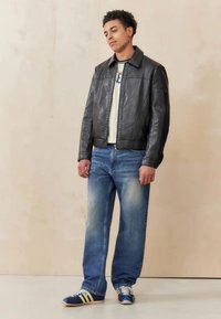 Black leather bomber jacket, light-colored t-shirt, light blue jeans, and blue sneakers with cream accents. Stands against a beige background.