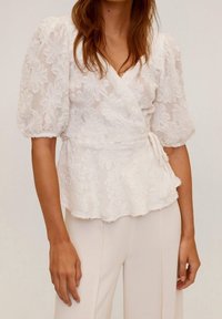 Top bianco avvolgente con elaborato pizzo floreale, maniche a sbuffo e orlo a peplo. Abbinato a pantaloni fitted di colore chiaro.