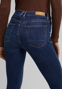 Jeans de mezclilla azul oscuro con forma ajustada, que presentan dos bolsillos traseros, costuras visibles y una etiqueta de cuero marrón claro en la cintura.