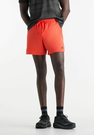 Person trägt leuchtend orangefarbene Sportshorts, schwarze und graue Laufschuhe sowie schwarze Socken und steht vor einem schlichten weißen Hintergrund.