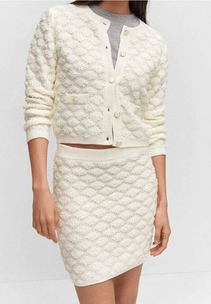 Femme portant un cardigan en maille texturée crème avec des boutons et une jupe taille haute assortie, chemise grise visible sous le cardigan.