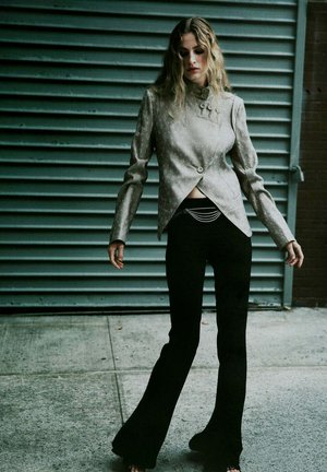 Mujer con pelo rubio ondulado lleva una chaqueta ajustada de color gris claro y pantalones negros acampanados, de pie en una acera frente a una persiana metálica.