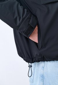 Persona que lleva una chaqueta negra con bucle ajustable y puño de Velcro, con la mano metida en el bolsillo lateral, combinada con jeans de mezclilla azul claro.