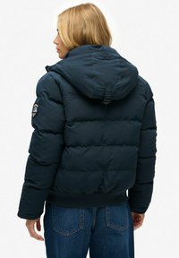 Chaqueta acolchada azul marino con capucha, que cuenta con secciones cosidas para aislamiento. Incluye un parche con el logo en el hombro izquierdo y un dobladillo elástico.