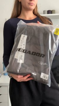 Μαύρο ένδυμα σε διαφανή πλαστική συσκευασία με το λογότυπο «PEGADOR». Το αντικείμενο εμφανίζει ετικέτες φροντίδας και γραμμικό κώδικα. Το σχήμα του αντικειμένου είναι ορθογώνιο, επίπεδο.