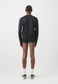 Sweat-shirt noir à manches longues, coupe ajustée, et poignets côtelés, associé à un short noir. Le modèle porte des chaussettes blanches et des baskets noires.