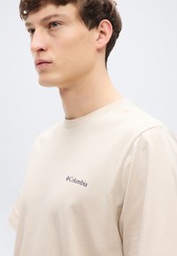 Μπεζ κοντομάνικο t-shirt από βαμβάκι, με στρογγυλή λαιμόκοψη και μικρό λογότυπο Columbia σε σκούρο μπλε στην αριστερή πλευρά του στήθους.