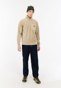 Beige fleece trui met een kwart-rits en borstzak, gecombineerd met donkerblauwe jeans en een groene gebreide beanie. Getextureerde stof en casual pasvorm.