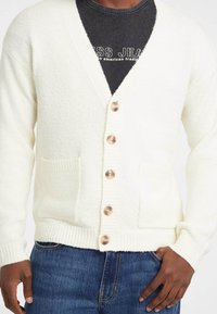 Witte gebreide cardigan met een v-halsontwerp, vijf houten knopen, ribgebreide manchetten en voorzakken, gecombineerd met een donker shirt en denim jeans.