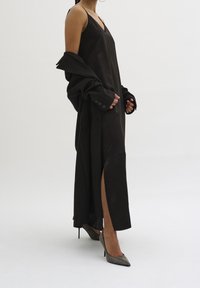 Robe fourreau en satin noir avec fente latérale, bretelles réglables et un manteau noir ample. Portée avec des escarpins métalliques à bout pointu. Texture lisse.