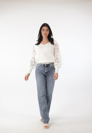 Vrouw met lang donker haar, gekleed in een witte trui met kanten mouwen, blauwe spijkerbroek en witte sandalen, staand tegen een effen witte achtergrond.