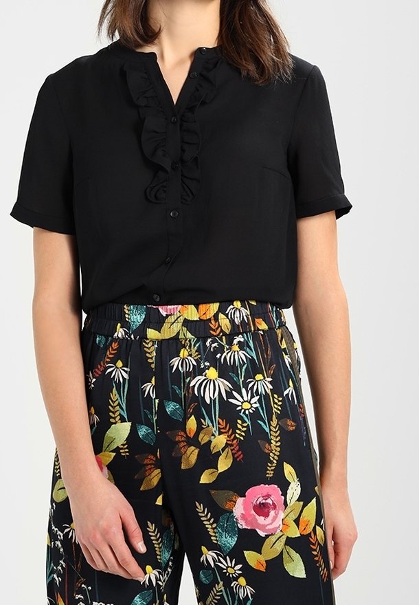 Femme portant un chemisier noir à manches courtes avec un détail volanté et un pantalon noir avec des motifs floraux colorés.