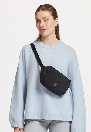 Donna con maglione azzurro chiaro e jeans grigi con una borsa a tracolla nera appoggiata sul petto su sfondo bianco.