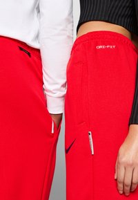Červené tepláky s označením Dri-FIT, skladajúce sa z vreciek na zips a loga Nike. Látka je hladká s jemným leskom; elastický pás.