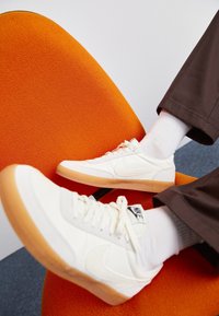 Białe sneakersy Nike z gumowymi podeszwami, noszone z białymi skarpetkami i ciemnymi spodniami, wypoczywają na pomarańczowym teksturowanym krześle.