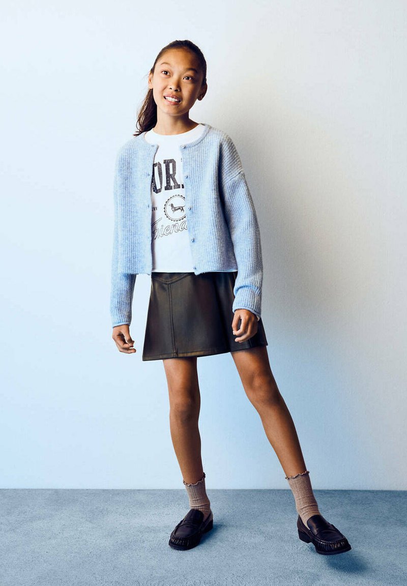Fille debout portant un cardigan bleu clair, un t-shirt blanc graphique, une jupe noire, des chaussettes beige et des chaussures noires sur un fond uni.