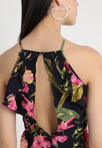 Hedvábné bezrukávové šaty mají tmavě modré pozadí s živými růžovými a zelenými tropickými vzory, volánkový zadní díl a otvor ve tvaru klíčové dírky.