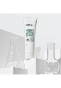 Tubo de concentrado ácido Redken bonding presentado junto a tres vasos de precipitados de vidrio transparente sobre plataformas geométricas blancas.