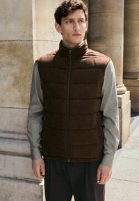 Brun puffervest med høj krave, lynlåse foran og sidelommer, parret med en lysegrå langærmet skjorte og mørke bukser.